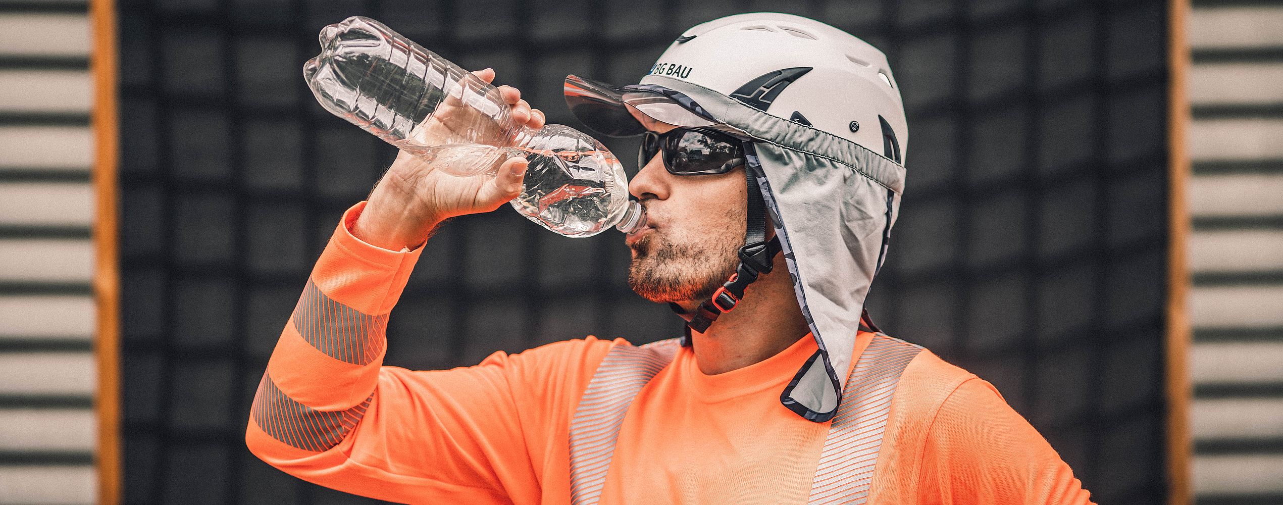 Ein Bauarbeiter mit Schutzhelm, Sonnenbrille und Nackentuch trinkt Wasser in der Pause.